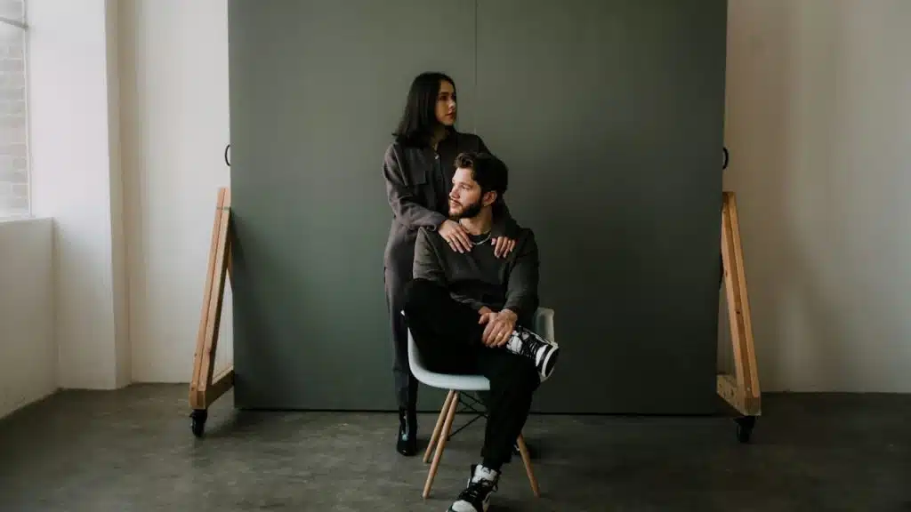 A woman resting her hands on a man’s shoulders as he sits on a chair while she sits on its armrest.