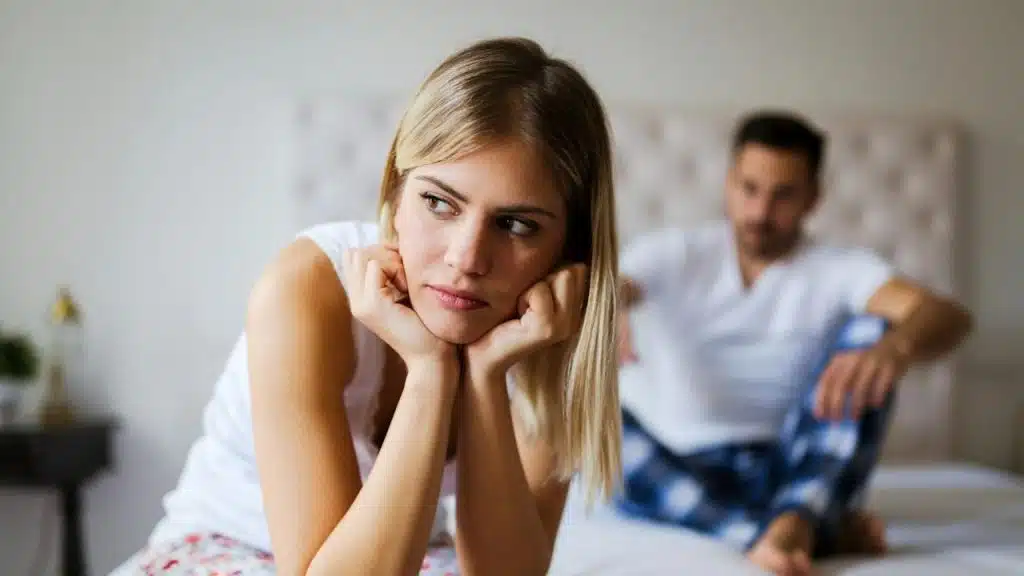 A woman holding her face in her hands and looking cross while a man sits on a bed in the background.