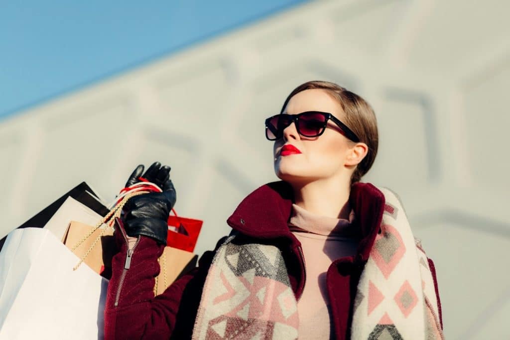 A woman wearing sunglasses holds shopping bags and looks upwards.