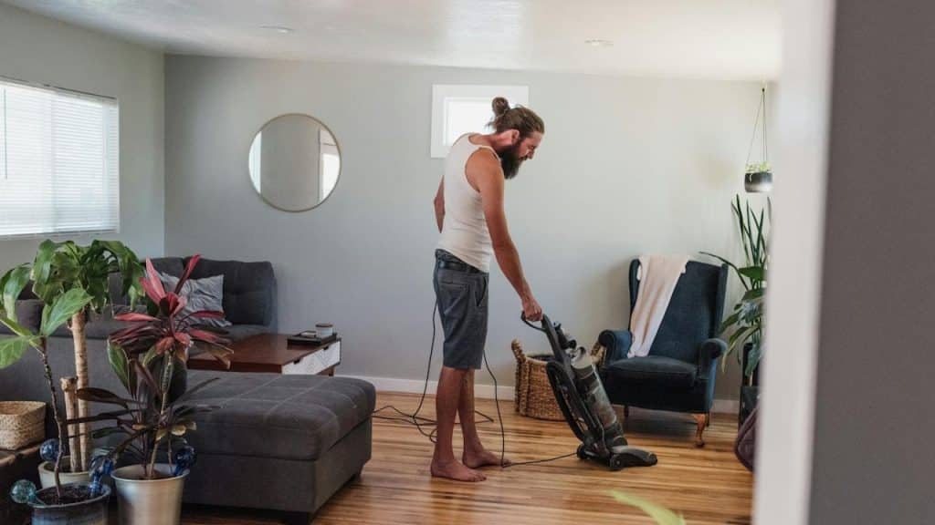 Man with long hair and a beard, wearing a white tank top, vacuuming a hardwood floor in a living room filled with plants.
