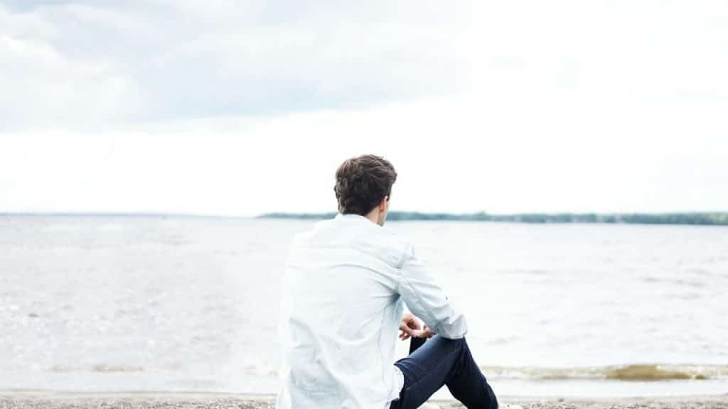 A man sitting by the shore looking out over a calm body of water under a cloudy sky.