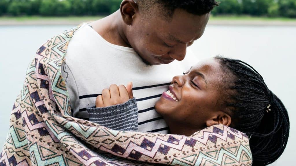 A couple wrapped in a blanket, smiling warmly while embracing outdoors near water.