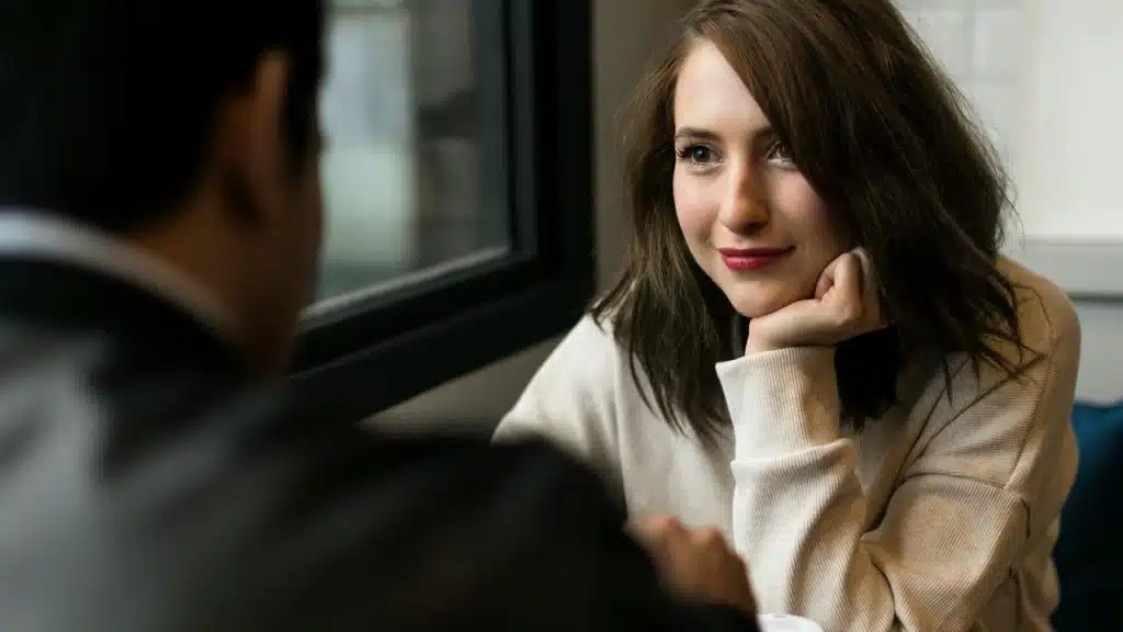 A woman resting her chin on her hand, smiling softly while listening to someone indoors.