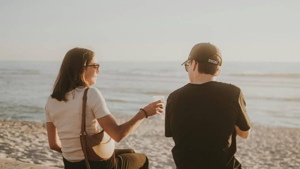 A couple sitting on a beach ledge, sharing a drink while looking out at the ocean.
