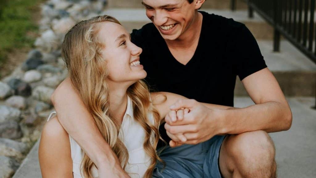 A smiling couple sitting close together outdoors, holding hands and laughing.
