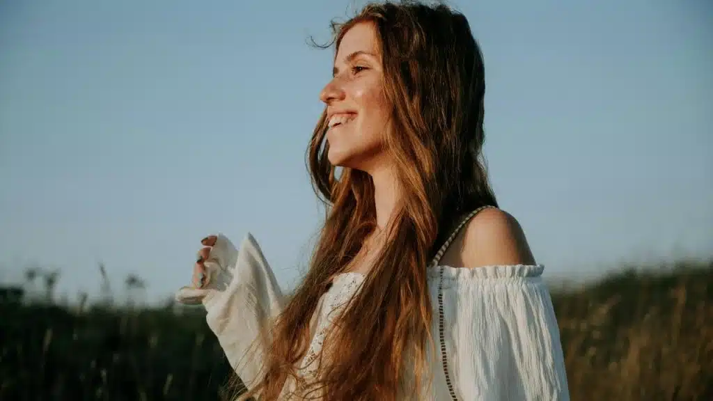 A smiling woman with long hair standing outdoors in warm sunlight.