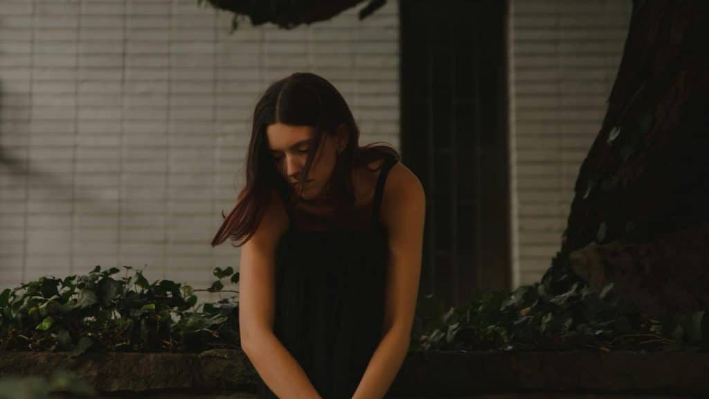 A woman sitting quietly outdoors with her head bowed near a tree.