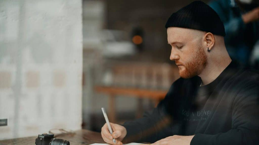 A man in a beanie writing at a table with a camera nearby.