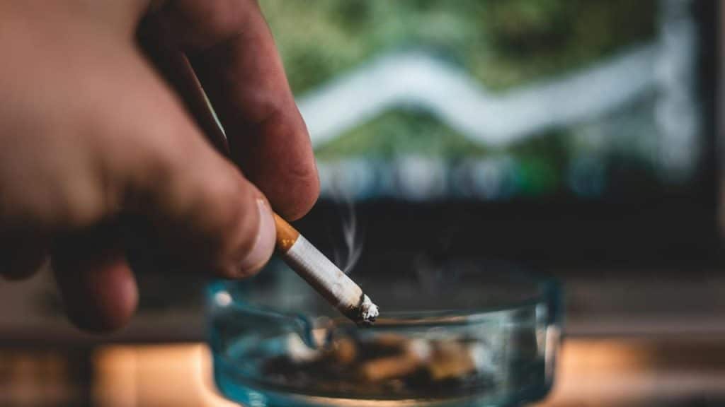 A hand holding a lit cigarette over an ashtray with a blurred line graph in the background.