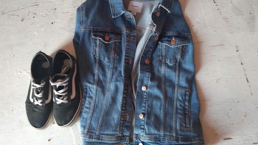 A denim jacket beside a pair of black sneakers on a light floor.