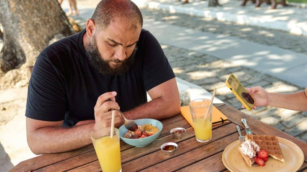 A bearded man eating a fruit bowl at a wooden table with glasses of juice and waffles nearby.