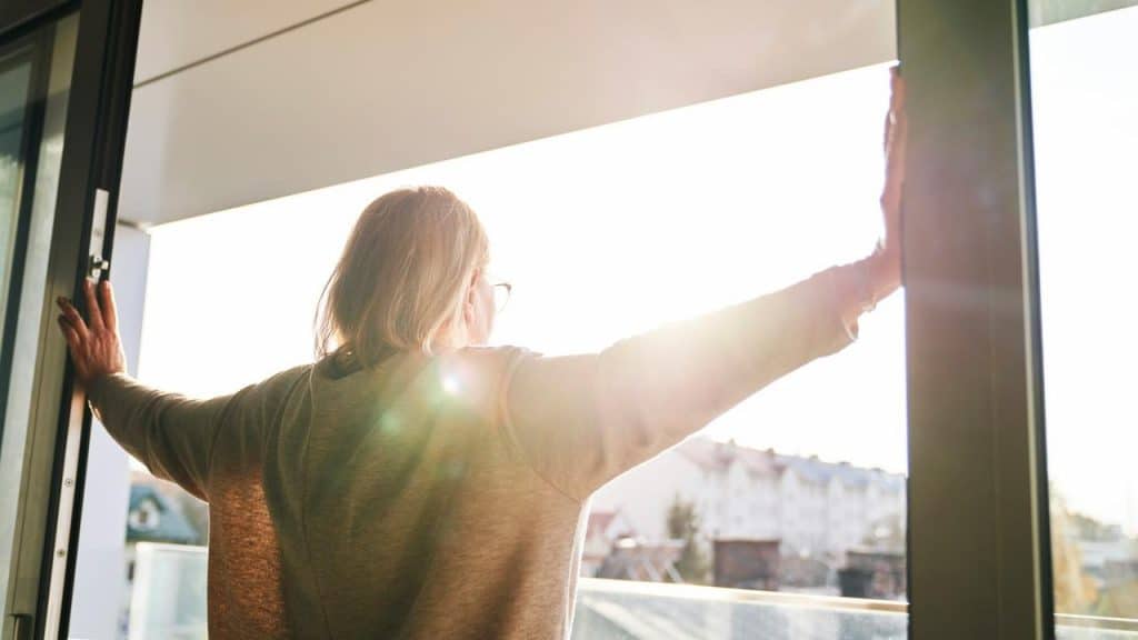 A person opening balcony doors and stretching arms toward the morning sunlight.
