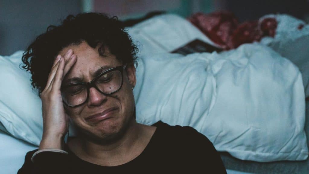 A person sitting on a bed with glasses, holding their head while crying.