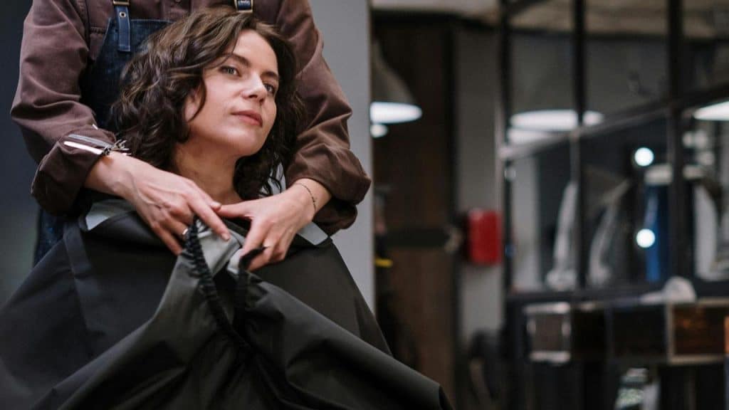 A woman sitting in a salon chair while a stylist adjusts a cape around her.
