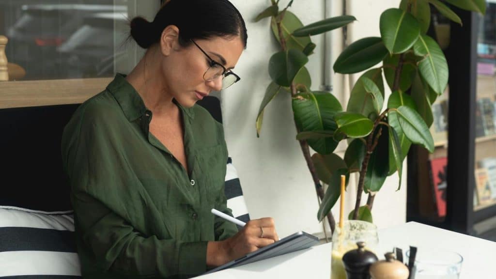 A woman wearing glasses writing on a tablet at a table near a plant.