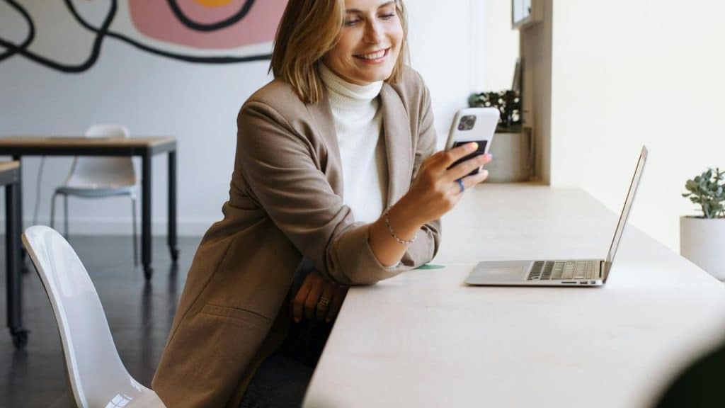 A woman smiling while looking at her phone beside a laptop at a table.