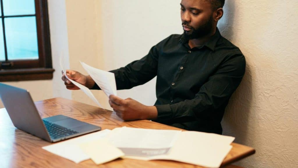 A man sitting at a desk reading papers beside a laptop.