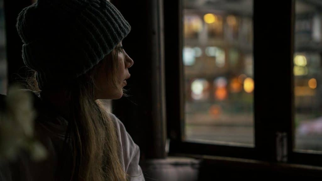 A person in a knit beanie gazing out a window at blurred city lights at night.
