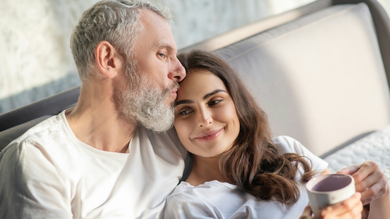 A bearded older man kissing a smiling younger woman’s forehead as she leans on him while having coffee.