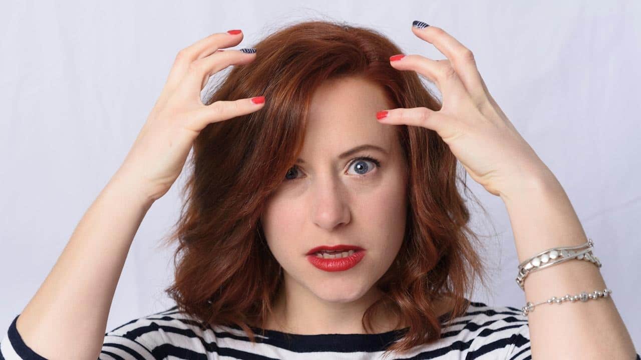 A woman with short red hair looks directly at the camera with a frustrated expression, raising both hands toward her head.