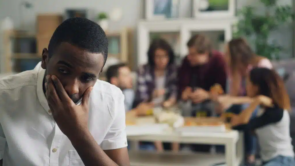 A man sits looking upset with a group of friends socializing in the background.