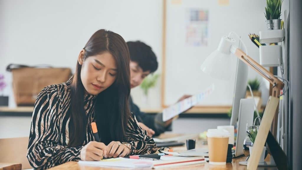 A woman is using a highlighter while working at the table with a man sitting next to her.