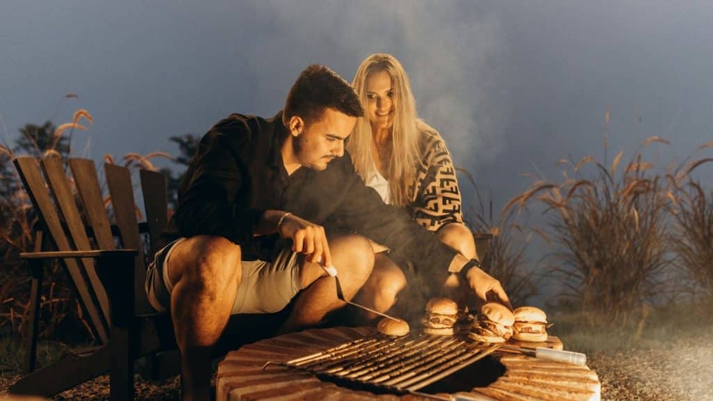 A man and woman grilling burgers in front of a bonfire.