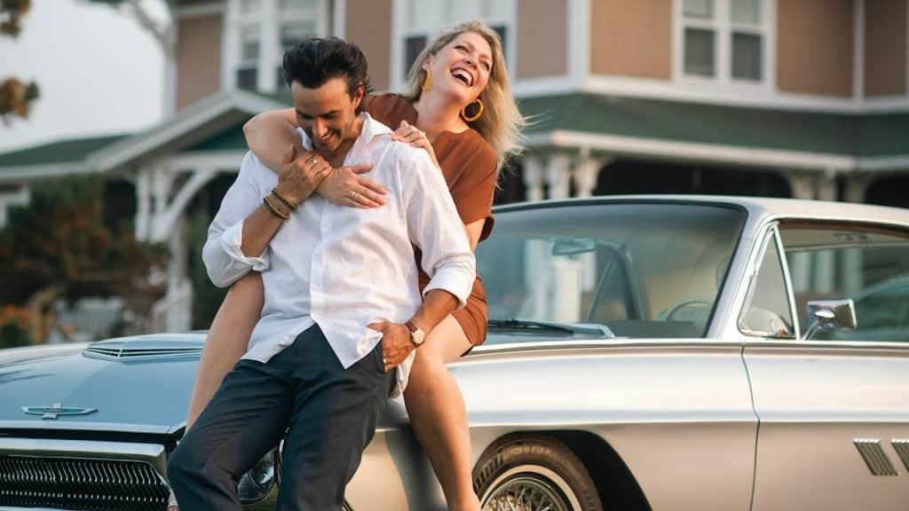 A man holding a woman’s hand who has her arms around his shoulders while laughing and sitting behind him on the car’s bonnet that he’s leaning against.