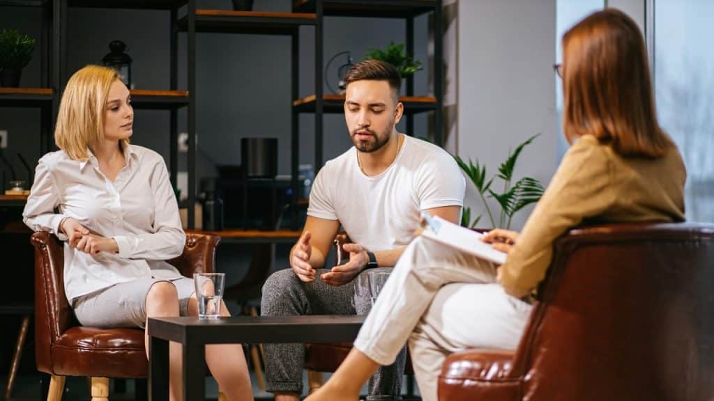 Man and woman sitting on leather chairs facing a person holding a clipboard in an office.