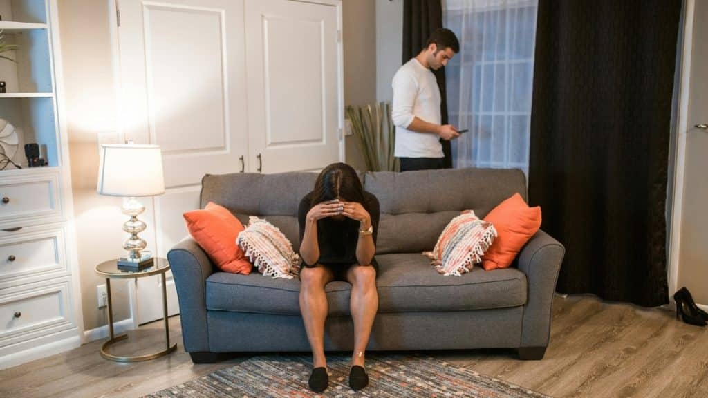 Woman sitting on a sofa with head in hands; man stands behind her looking at phone.