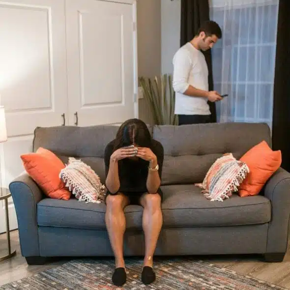 Woman sitting on a sofa with head in hands; man stands behind her looking at phone.