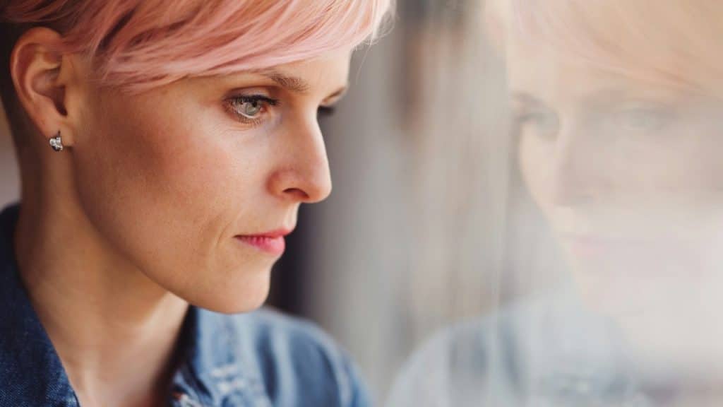 Close-up profile of a person with pink hair looking out a window with a soft reflection.