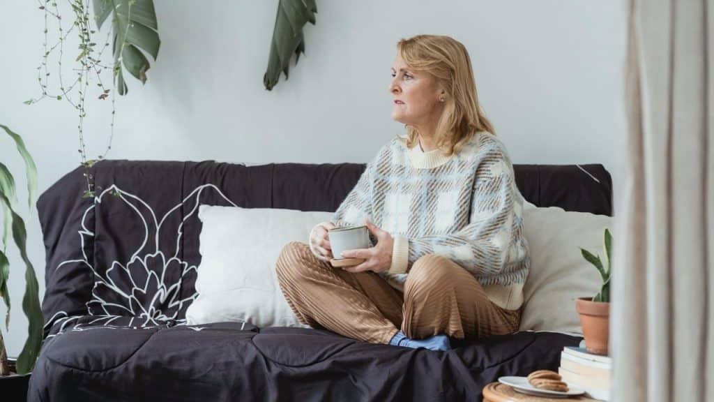 Woman sitting cross-legged on a sofa holding a mug and looking toward a bright window.