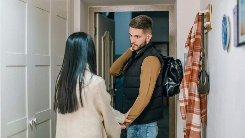 Man with a duffel bag standing in a doorway looking back at woman holding his arm.