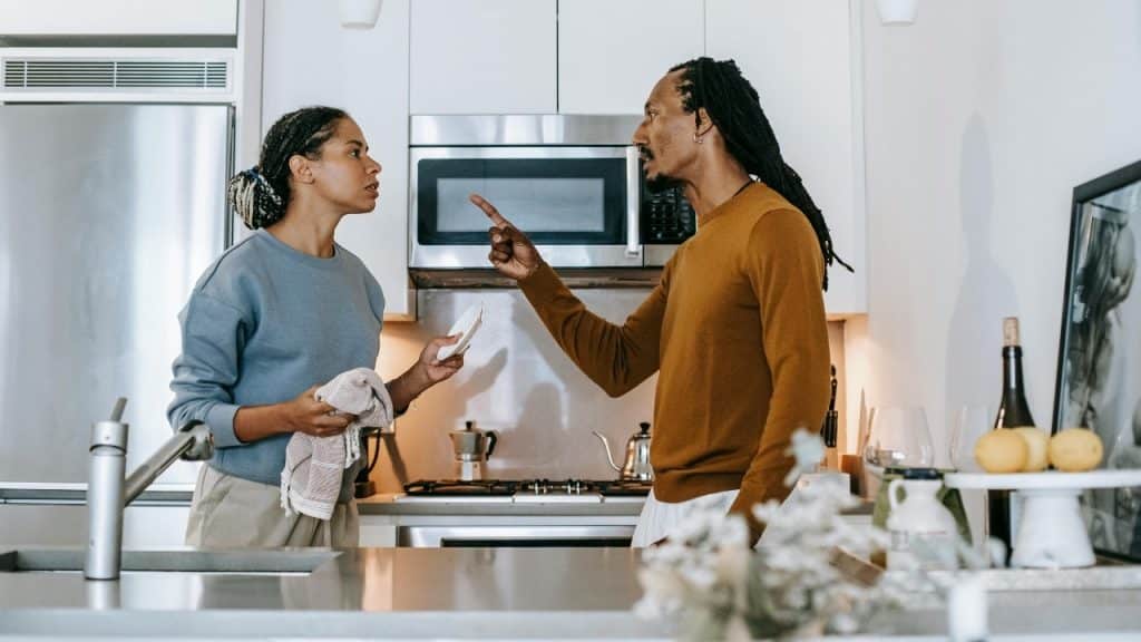 Man pointing a finger at a woman in a kitchen; both have serious facial expressions.