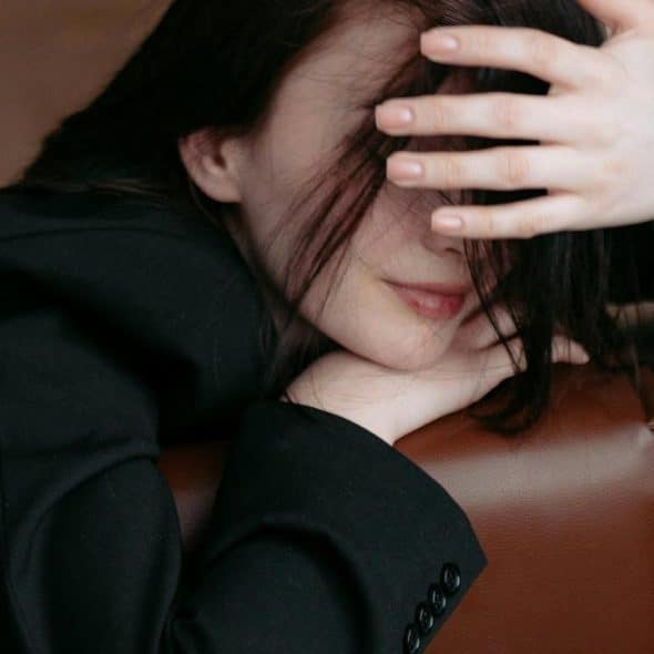 A woman in a black blazer resting her head on a chair with her hand partially covering her face.