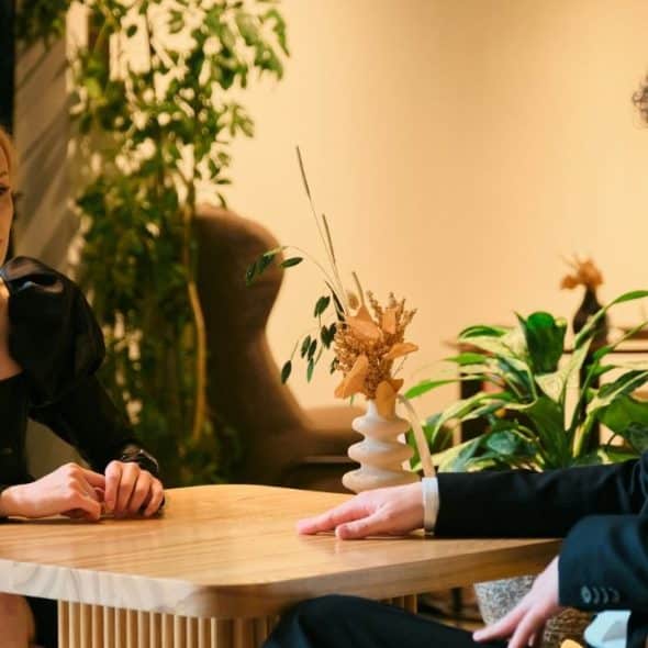 A blonde woman in a black dress sits at a wooden table facing a man.