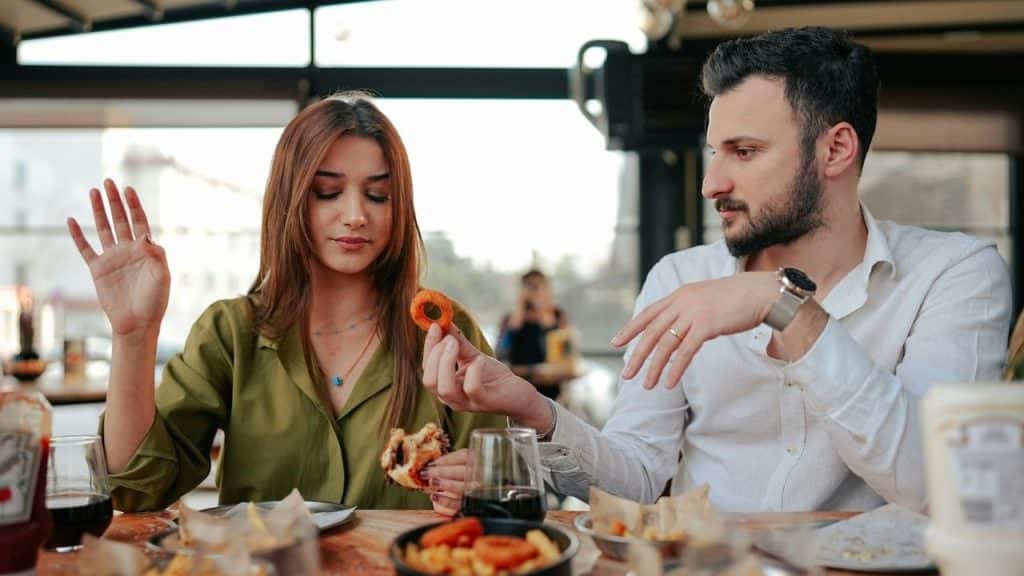 A man in a white shirt holds a food item toward a woman at a restaurant.