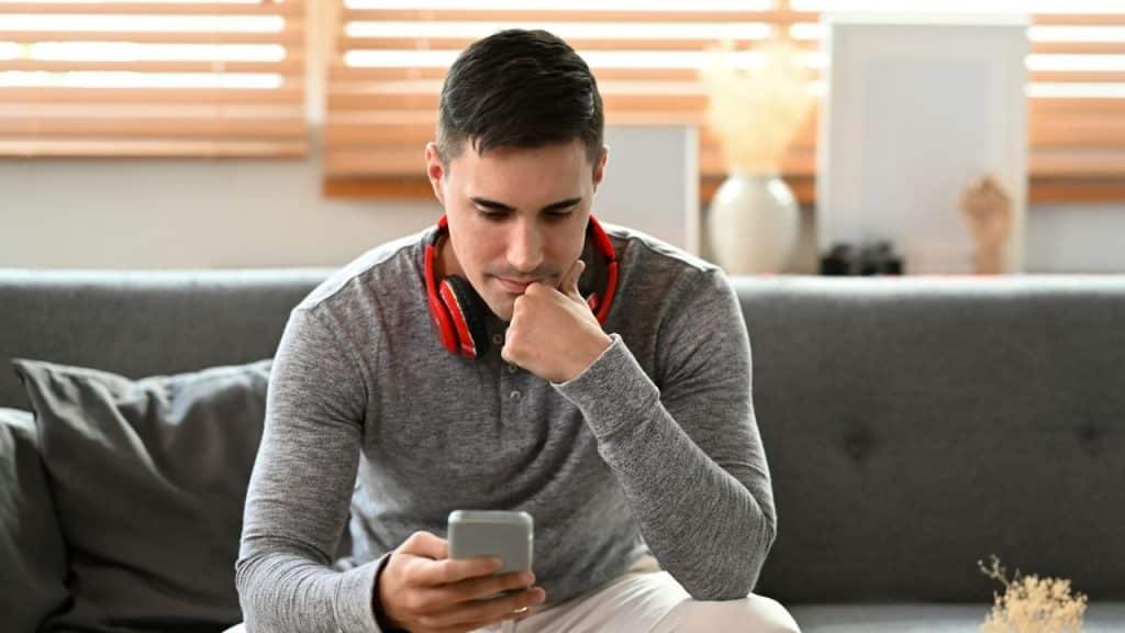 A man with red headphones around his neck looks down at his phone while sitting alone.