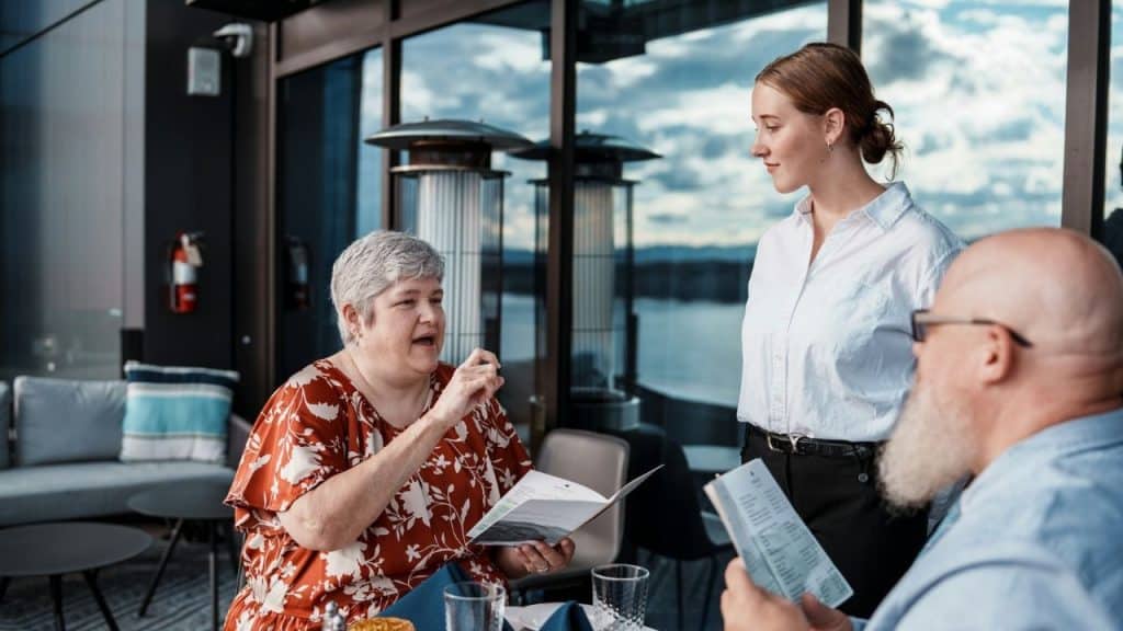 A woman in an orange floral top speaks to a server standing beside a restaurant table.