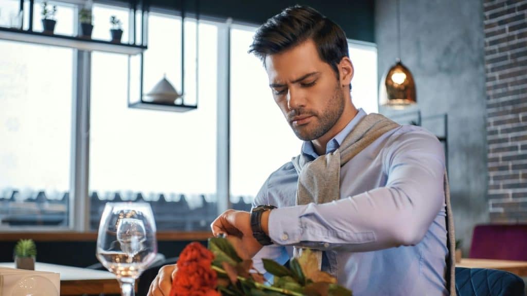A man in a blue shirt and grey sweater looks at his watch while sitting indoors.