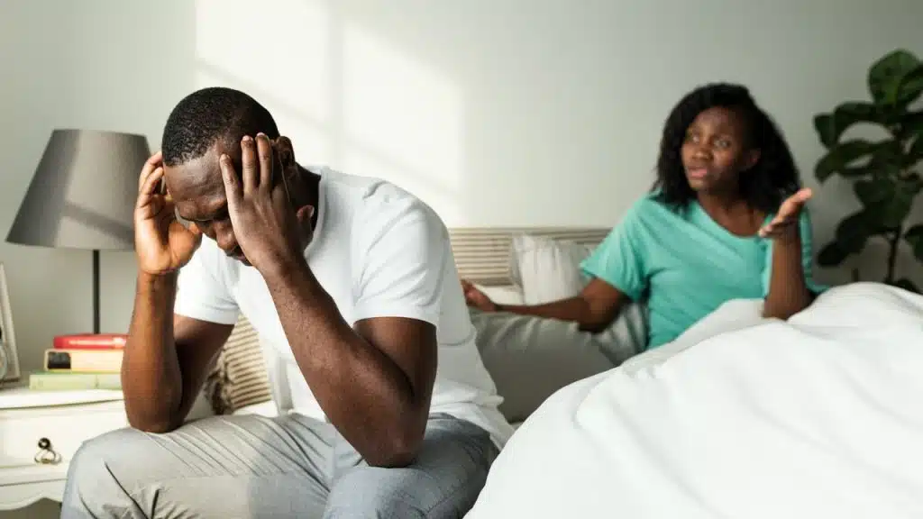 A man sits on a bed holding his head while a woman gestures in the background.
