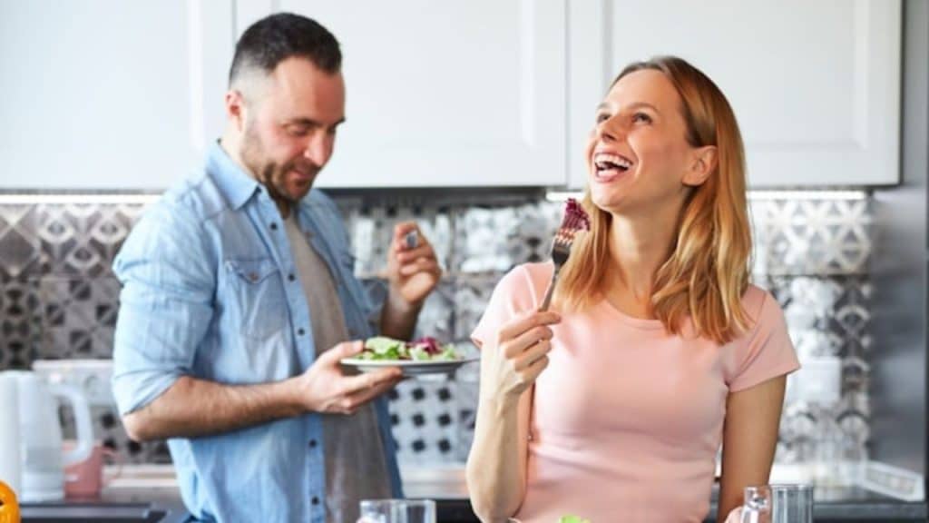 A woman in a pink shirt laughs while a man in the background holds a plate.