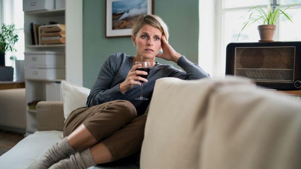 A woman sits on a couch holding a glass of dark liquid with a worried expression.