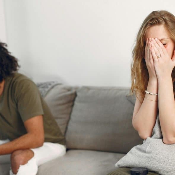 A woman sits on a sofa covering her face while a man sits looking away.