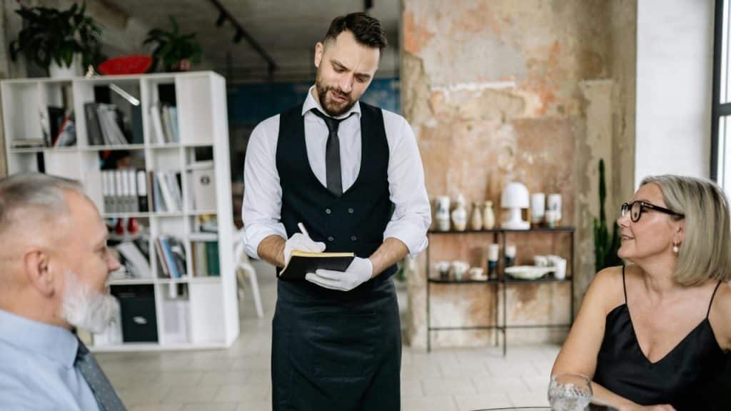 A server in a vest takes notes while standing between two people at a table.