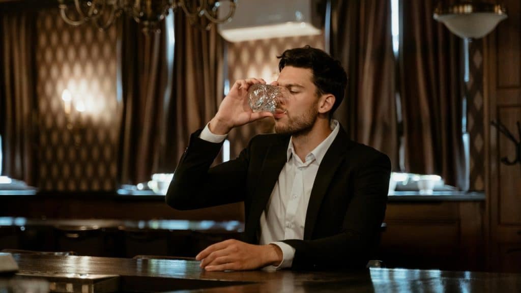 A man in a suit sits at a dark bar drinking from a crystal glass.