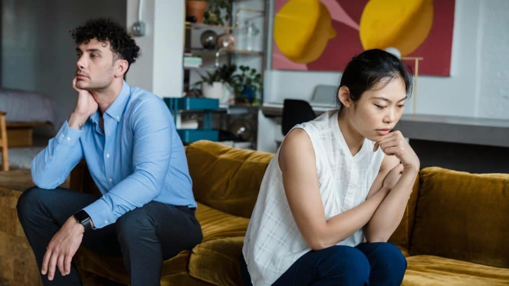 A man and a woman sit back-to-back on a yellow sofa with their heads resting on hands.