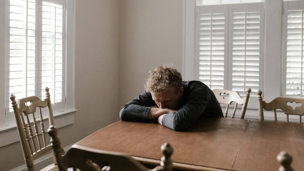 A man with curly hair rests his head on his arms at a wooden dining table.