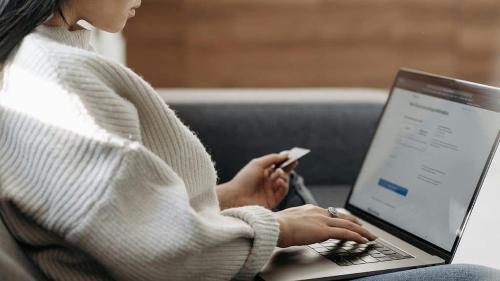 A woman sits on a couch using a laptop while holding a credit card in hand.